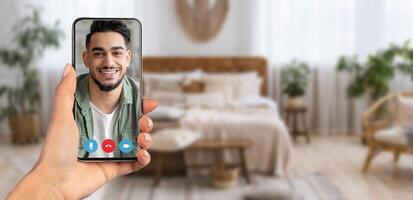 A person holds a smartphone displaying a smiling individual during a call from their cozy home interior. The background features a warm, inviting bedroom environment, emphasizing connection. photo