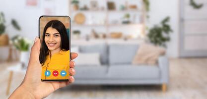 A person holds a smartphone showing a smiling individual on a call. The setting is a cozy living room with a sofa and decorative items visible in the background. photo