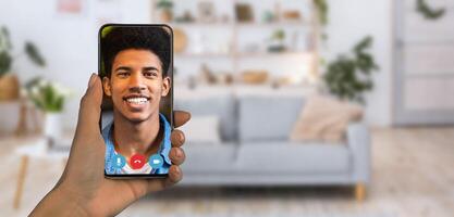 A person is holding a smartphone displaying a smiling individual during an online communication session. The cozy home setting features a stylish couch and decorative plants. photo