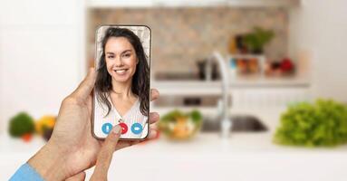 A person is using a smartphone to connect on a call while in a kitchen setting. The hand displays a friendly gesture on the screen, which shows a smiling participant. photo