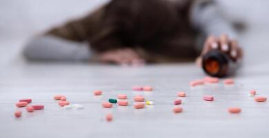 Young woman committing suicide, lying on floor with tablets scattered from jar, selective focus. Depression and drugs overuse, mental illness or psychological disorder concept photo
