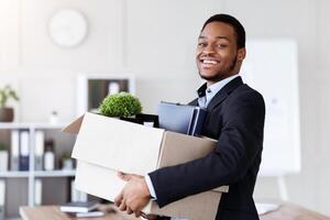 Happy black businessman moving to new office with box and his belongings, copy space. Successful african american manager holding box with working stuff, changing working place photo