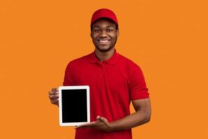 Delivery Concept. Portrait of smiling black male courier holding digital tablet with empty screen for mock up and showing to camera, wearing red cap and uniform, standing isolated on orange background photo