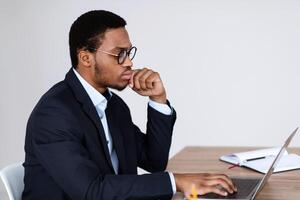 Pensive african american businessman in glasses working with laptop in office, looking at computer screen and touching his face, looking for creative solution or new clients, side view, copy space photo