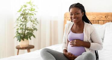 Maternity Concept. Portrait Of Happy Expectant Black Lady Posing On Bed At Home, Embracing Her Pregnant Belly And Smiling At Camera, Positive Awaiting Woman Enjoying Her Healthy Pregnancy, Free Space photo