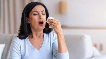 Sick mature woman sneezing and coughing, holding tissue paper napkin, sitting on the couch at home. Brunette ill middle-aged lady got cold or flu, having fever and runny nose, copy space photo