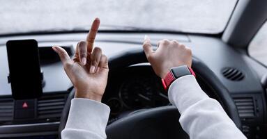Traffic problems, violation of rules, anger and quarrel. Upset millennial african american lady controls steering wheel with her hand and shows middle finger, view of window with drops and city road photo