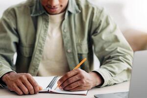 Remote learning concept. Black student writing in notebook during online lesson at home, focus on hands. Cropped view of African American youth participating in webinar, taking educational course photo