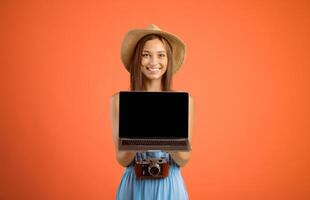 Happy young woman traveller holding laptop with blank screen over yellow studio background, website for travellers and tourists concept. Smiling lady tourist showing empty screen on her notebook photo