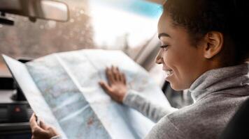 Young woman travel alone with map in car in new town. Smiling beautiful millennial african american lady driver looking at chart and searching for way or planning route for journey, empty space photo