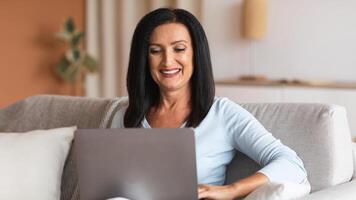 Domestic Comfort. Portrait Of Positive Mature Woman Using And Working On Laptop At Home, Enjoying Free Time, Smiling Lady Sitting On The Couch With Computer On Her Lap, Watching photo