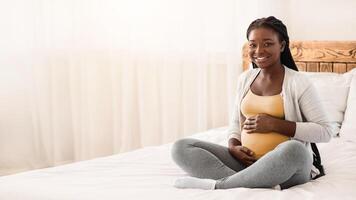 Pregnancy Time. Happy Black Pregnant Woman Sitting On Bed Touching Belly, Young Expectant Lady Posing In White Bedroom, Cuddling Tummy And Smiling At Camera, Resting At Home, Copy Space photo