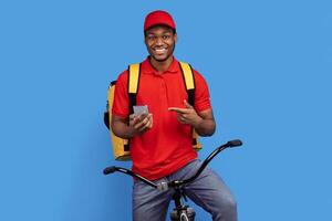Courier Service Application. Smiling black delivery guy using smartphone and pointing at it, wearing red uniform, cap and thermo bag, riding bicycle, isolated on blue studio background photo