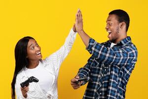 Victory together, love, friendship and modern entertainment. Joyful excited zoomers african american male and female give high five with gamepads in hands, isolated on yellow background, studio shot photo