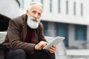 Modern aged man sitting on bench by business center and using digital tablet, websurfing or checking social media, copy space. Elderly businessman with long grey beard reading on pad photo