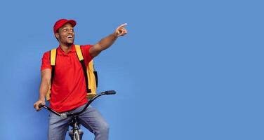 Look Here. Emotional surprised black delivery man in red uniform riding bicycle, wearing thermo backpack bag and pointing away at copy space isolated on blue studio background, banner, panorama photo