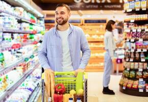 Smiling Cheerful Bearded Man Walking With Shopping Trolley Cart Along The Shelves In Grocery Store. Happy Guy Buying Groceries In Supermarket, Looking At Fridge With Dairy Products photo