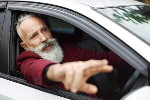Nervous senior man driver fighting on the road, stuck in traffic, side view. Stressed aged grey-haired businessman yelling and gesturing while driving car, going for business meeting, free space photo