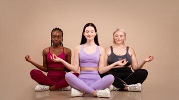 Beautiful body positive women of different size and appearances meditating together indoors, young happy multiethnic females in sportswear sitting in lotus pose with eyes closed over beige background photo