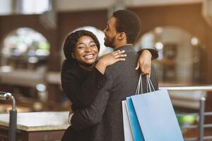 Free time and romantic date of african american couple at mall. Happy guy and lady in coats hugging and holding colored bags with many purchases during season sale and Black Friday, free space photo