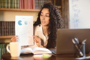 Portrait of female financial analyst having call on laptop, giving virtual presentation. Woman showing statistics, graphs and charts to camera, holding document paper, explaining report photo