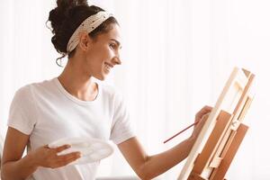 Young smiling woman artist painting at home, using easel, palette and paintbrush, side view, copy space. Closeup of creative lady drawing while having free time during quarantine photo