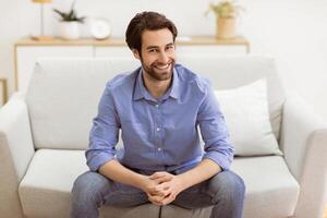 Cheerful Bearded Guy Sitting On Sofa Smiling To Camera Posing At Home. Male Portrait Of Successful Millennial Guy In Living Room Indoors. Confidence, People, Modern Lifestyle photo