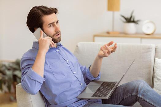 Discontented Guy Frowning Talking On Cellphone Sitting With Laptop, Having Computer Or Internet Connection Problem At Home. Displeased Customer Calling Hotline. Remote Work Issue. Selective Focus photo
