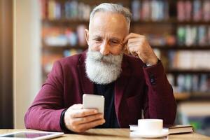 Senior grey-haired man with long beard drinking coffee at cafe, using smartphone, planning his day, copy space. Handsome aged businessman having coffee break, holding mobile phone photo