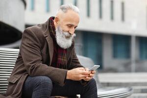 Happy elderly grey-haired man using smartphone while resting on street, sitting on bench and using mobile application, copy space. Handsome senior man using smartphone outdoors photo