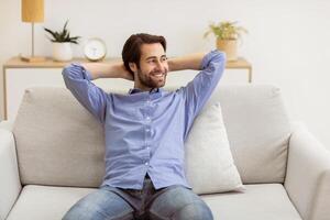 Relaxed Guy Sitting Holding Hands Behind Head On Sofa Relaxing At Home. Pleased Man Resting In Living Room Looking Aside. Weekend, Finally. Domestic Relaxation Concept photo