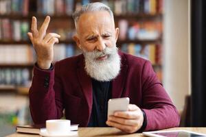 Angry senior man with grey hair and long beard using mobile phone while drinkin coffee at cafe, having negative face expression and gesturing, got bad news, having digital tablet by him, copy space photo