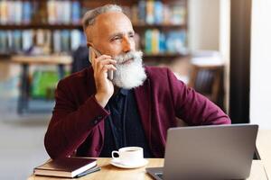 Busy senior businessman talking on mobile phone while drinking coffee at cafe, using laptop, looking at copy space. Elderly handsome bearded man in suit working online, using computer photo