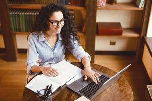 High angle view portrait of smiling young woman sitting at desk, typing on keyboard and using laptop computer. Lady watching online tutorial, webinar or seminar, studying at home, working remotely photo