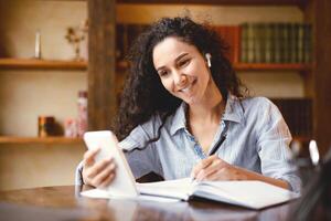 Remote Education. Smiling female student in wireless earphones sitting at desk, using smartphone and writing in notebook, taking notes, watching lesson, lecture or webinar, studying online at home photo