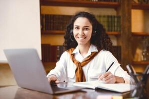 E-learning And Distant Education During Quarantine. Positive young female student in headset studying online, using laptop, writing and taking notes in her notebook, looking and posing at camera photo