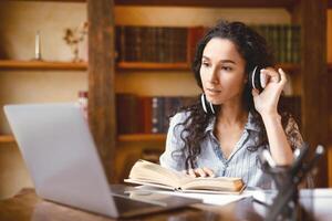 Distance Education. Portrait of woman touching wireless headset sitting at desk, using laptop and reading book. Lady watching tutorial, lecture or webinar, studying online at home. E-learning photo