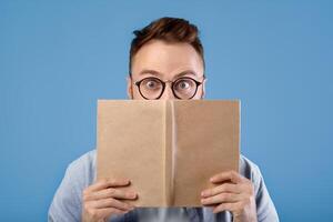 Shocked young guy in glasses reading book, opening his eyes wide in surprise on blue studio background. Puzzled reader stuck in textbook, finding exciting information, learning something interesting photo