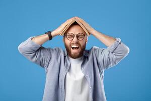 Furious young man putting book on his head and screaming on blue studio background. Bearded millennial guy tired of studying, cannot do difficult home assignment or meet deadline photo