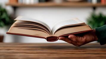 A hand holding an open book over a wooden table with blurred plants in the background photo