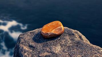 A polished orange stone resting on a rugged rock by the ocean, waves crashing in the background photo