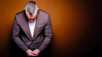 A well-dressed man in a suit bows his head against a warm, gradient background, conveying contemplation photo