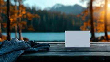 Blank card on wooden table with cozy blanket by serene lake and autumn trees in background photo