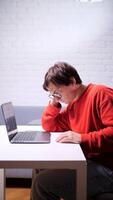 uma cansado homem dentro uma vermelho suéter é Deslizamento às uma mesa com uma computador portátil video