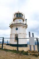 Historic lighthouse stands on rocky coast under cloudy sky with surrounding fence and lookout equipment photo