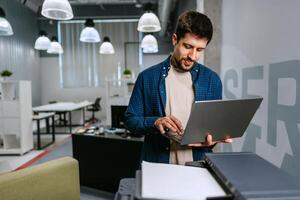 Man working on laptop in modern office setting during daytime while using printer photo