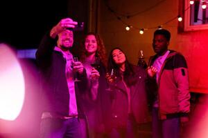 Friends enjoy a lively evening gathering while taking a selfie with drinks in a warmly lit outdoor space photo