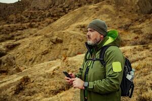 Man standing on rocky terrain in rugged outdoor gear while checking his phone during an adventure in nature photo