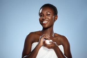 Smiling woman holding skin cream while wrapped in towel against a light blue background photo