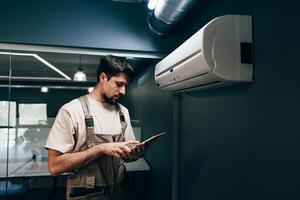 Technician checks settings on tablet while performing maintenance on air conditioning unit in modern office photo